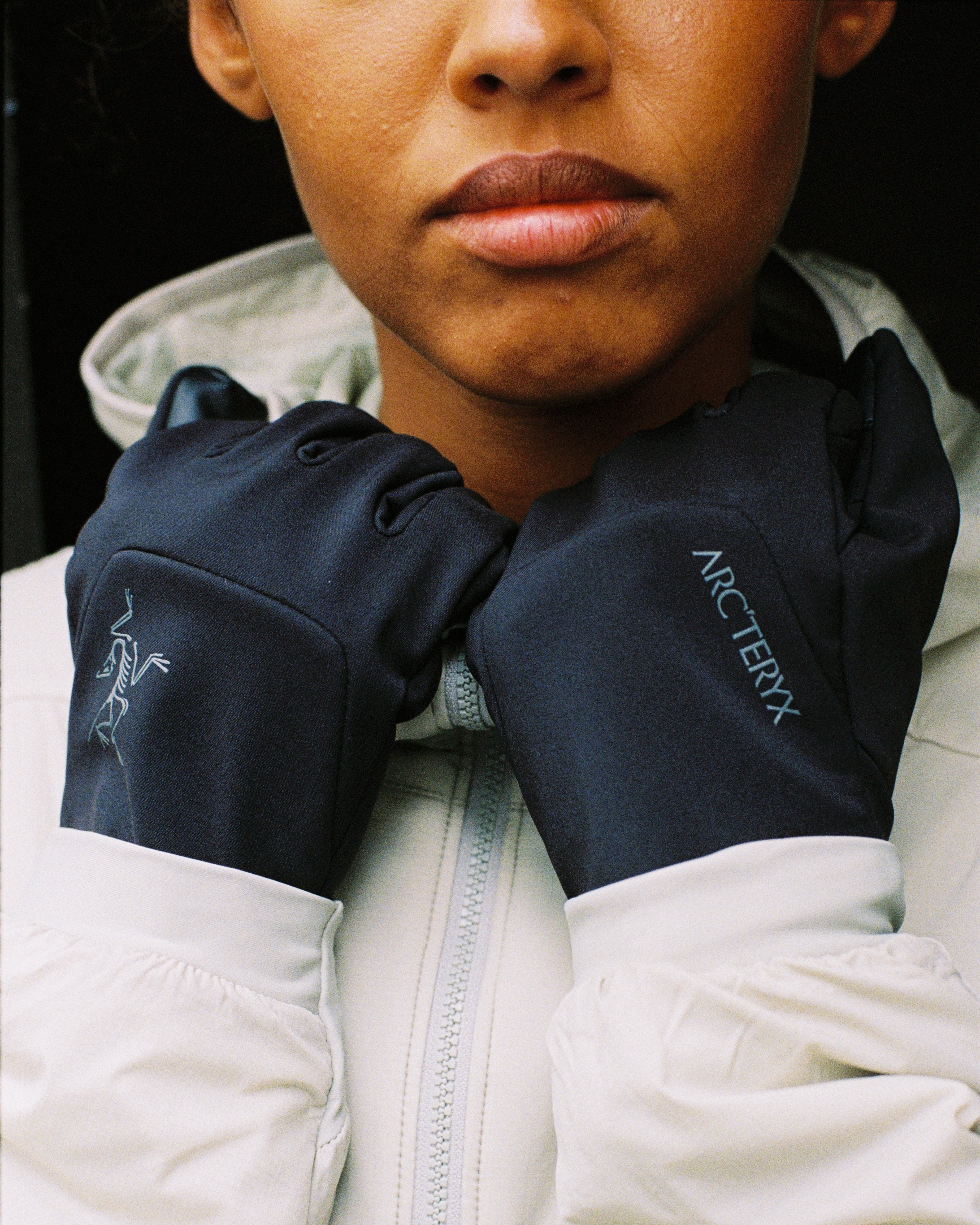 A close-up of a person wearing black Arc'teryx gloves over a white zip-up jacket, with the Arc'teryx logo visible on the gloves.