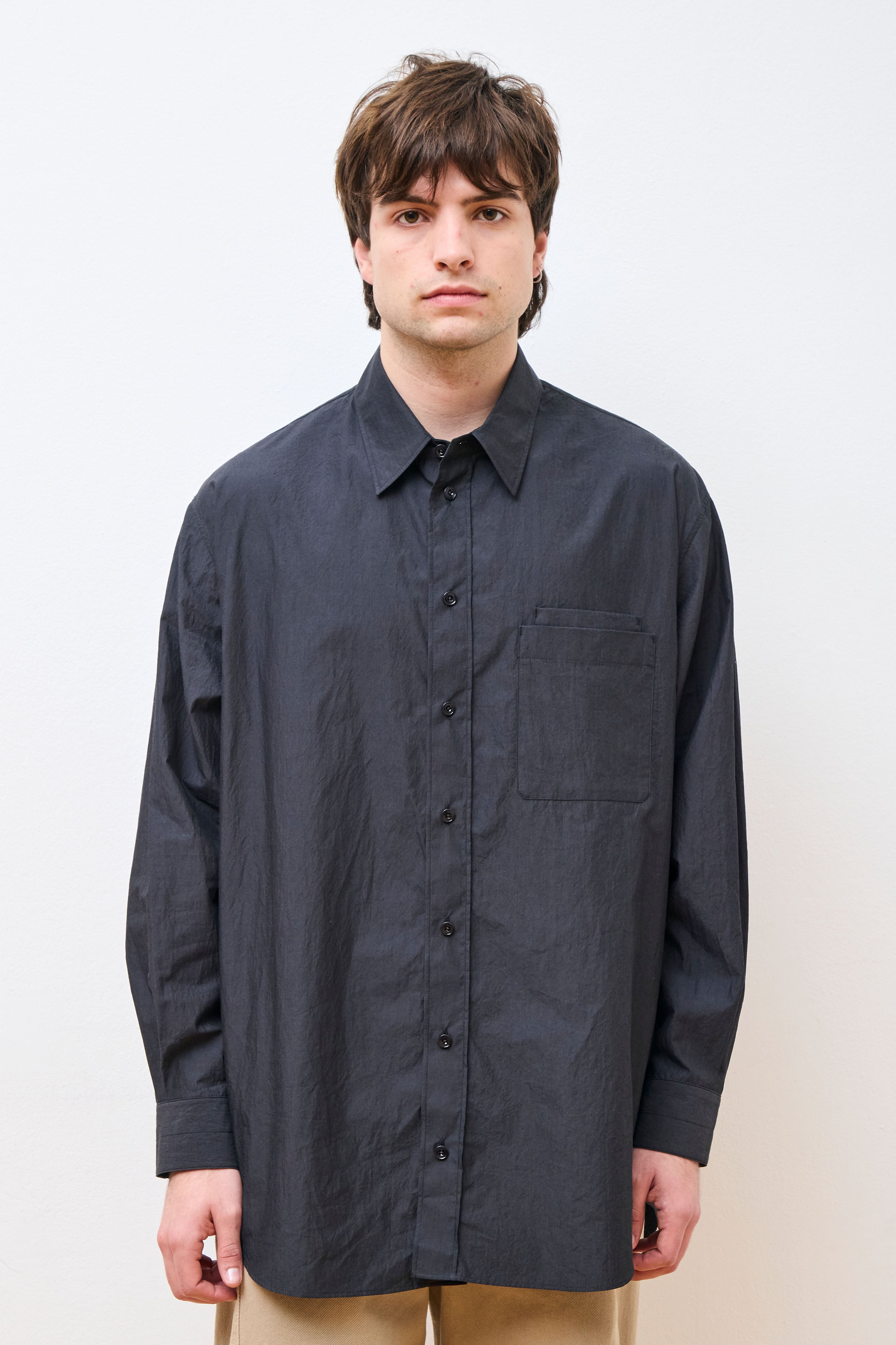 A male model wearing a long-sleeved, button-up, charcoal-colored shirt with a pointed collar and a double chest pocket, paired with light beige trousers.
