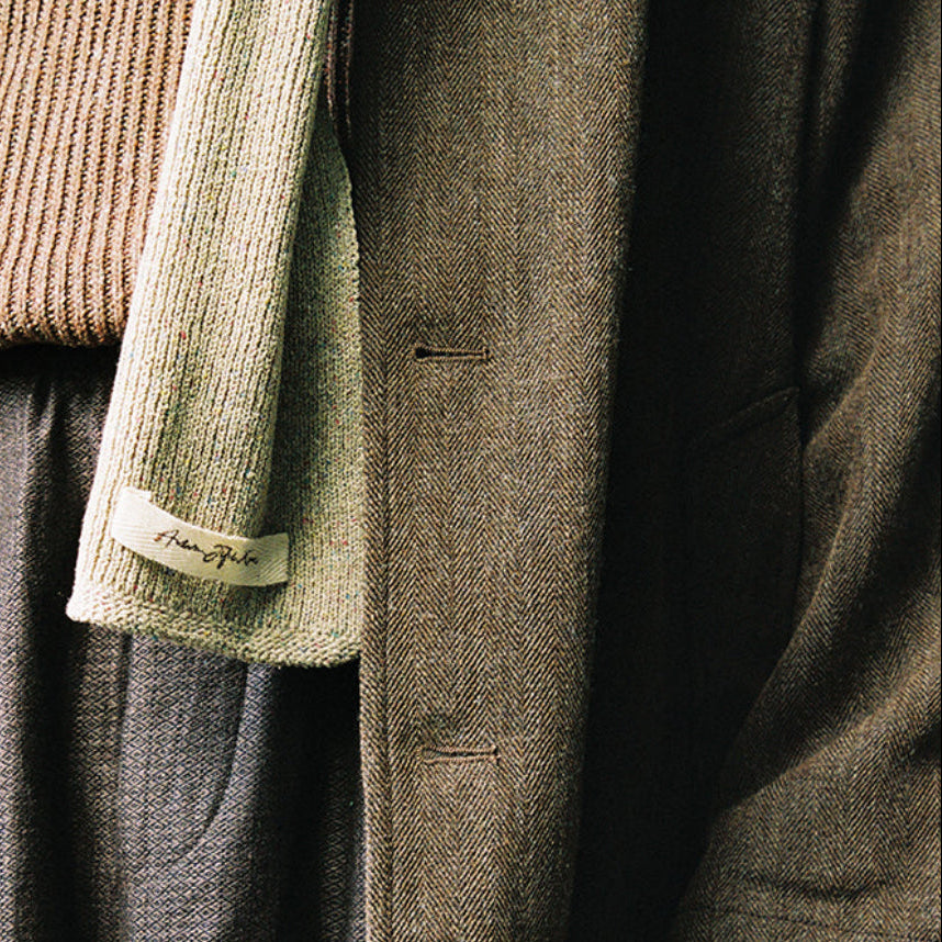 A close-up of a person wearing layered clothing, including a taupe knit sweater, a light sage green cardigan draped over the shoulder, dark gray textured trousers, and a dark brown coat. A small fabric tag is visible on the cardigan sleeve.