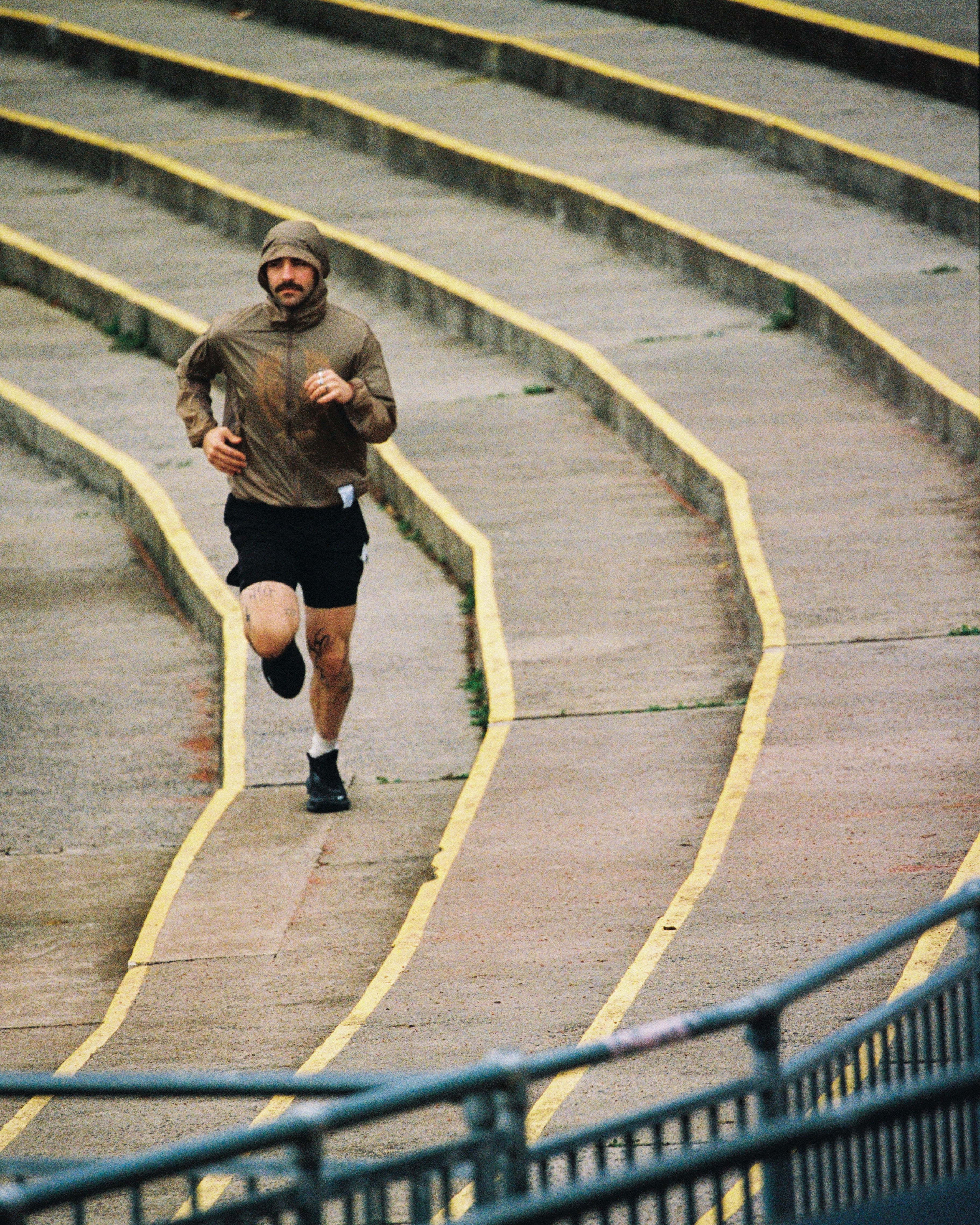 A man wearing a brown hooded jacket and black shorts is running up curved concrete stadium steps with yellow painted lines.
