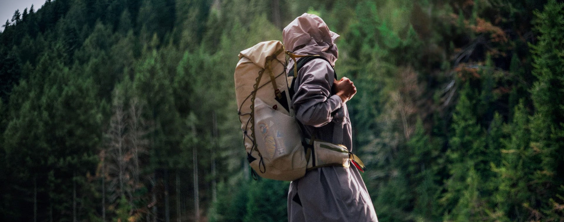 A person hiking through a dense pine forest, wearing a long gray hooded raincoat and carrying a large beige backpack.