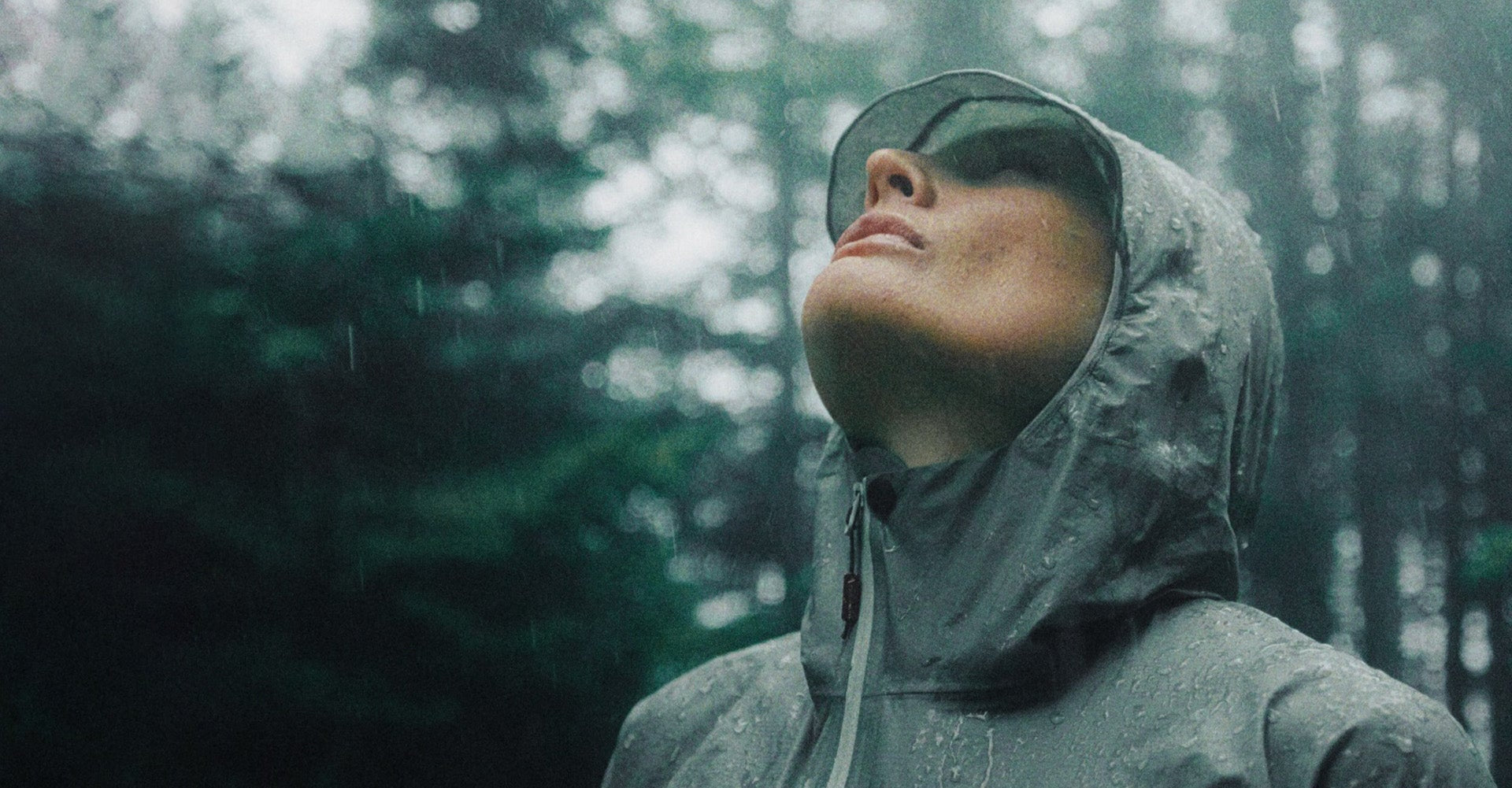 A person wearing a dark gray waterproof hooded rain jacket and matching visor, standing in the rain outdoors with a blurred forest background.